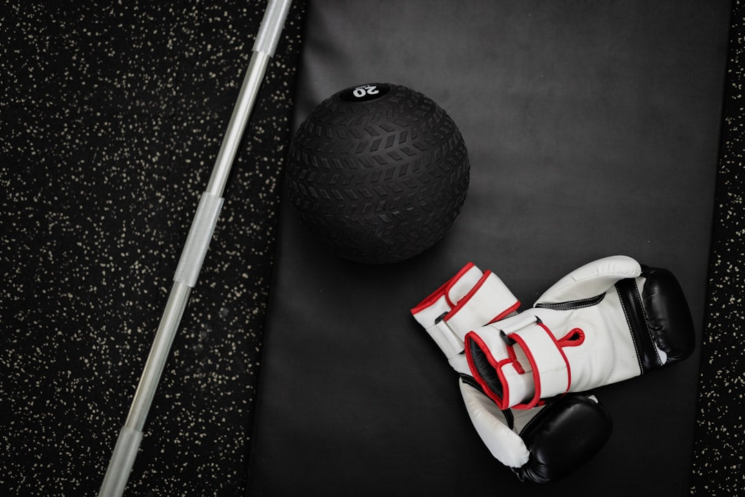 A high-angle shot of various workout equipment on a black mat and black speckled gym floor, including a black 20-pound slam ball, a pair of white and black boxing gloves with red trim, and a long, silver fitness bar.