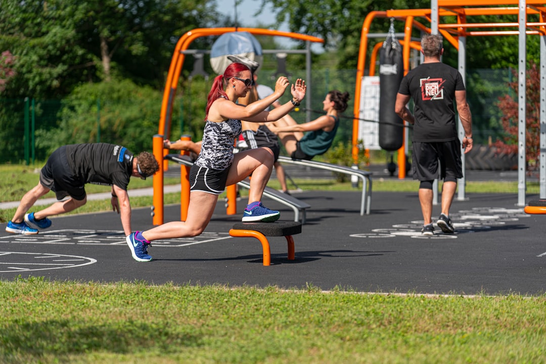 People exercising outdoors in a fitness area with orange metal equipment; a woman is performing a lunge on a step platform in the foreground.