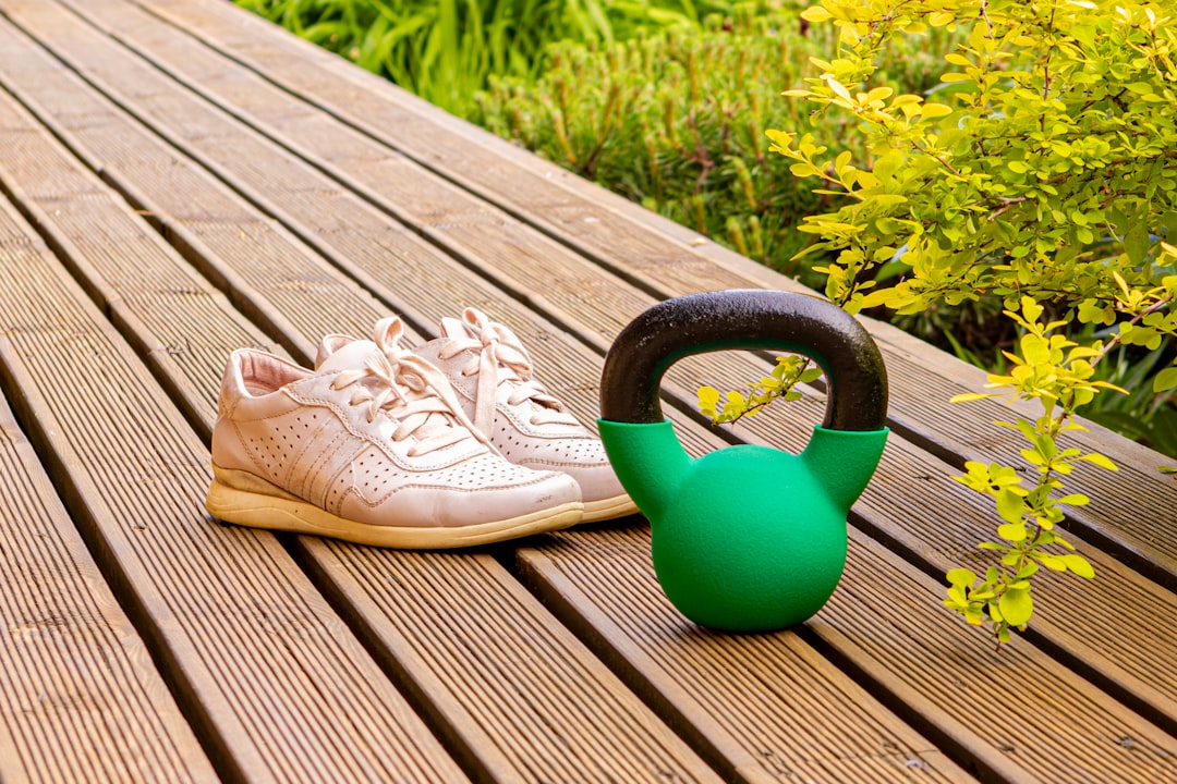A pair of beige athletic shoes and a green kettlebell placed on a wooden deck surrounded by greenery, symbolizing outdoor fitness or home workout setup.